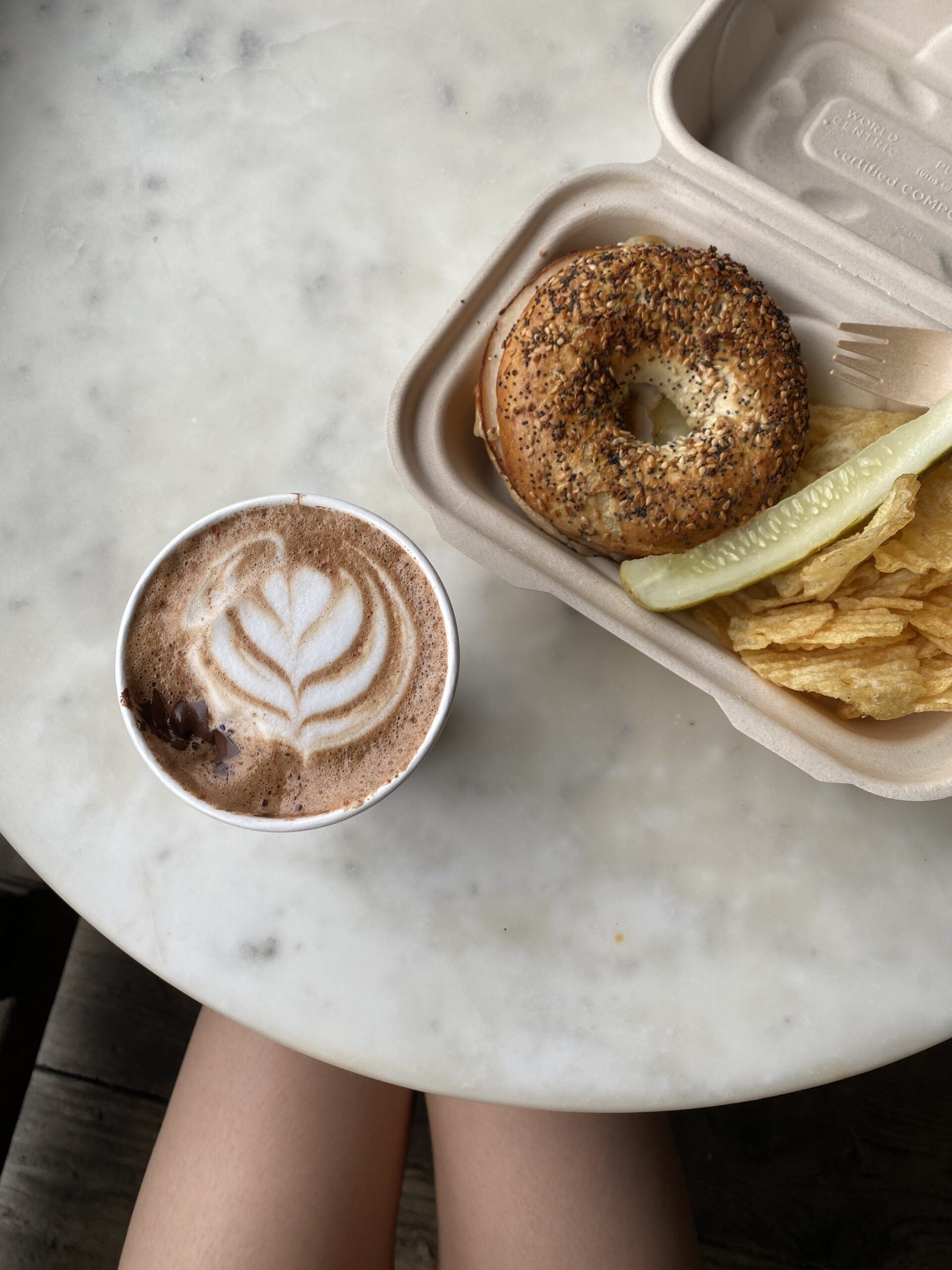 A delightful Chocolate Mayan Mocha, bagel, pickle, and ruffled potato chips neatly set on a white marble table.